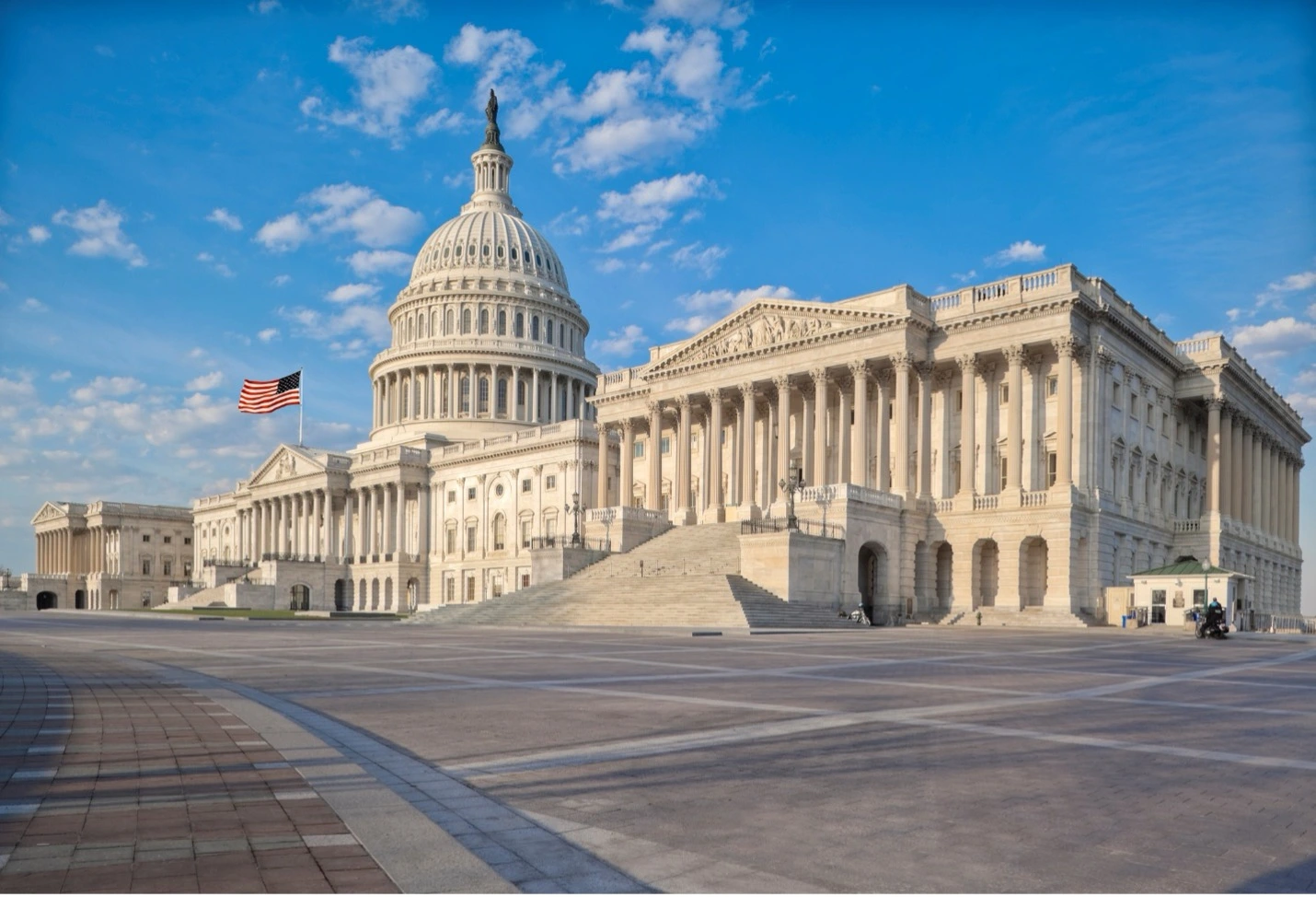 The United States' Capitol Hill with an American flag waving in the wind and partly-cloudy blue skies behind it